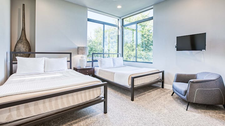 Amplia habitación con dos camas tamaño queen, con baño privado, grandes ventanales, un televisor montado en la pared y una decoración moderna.