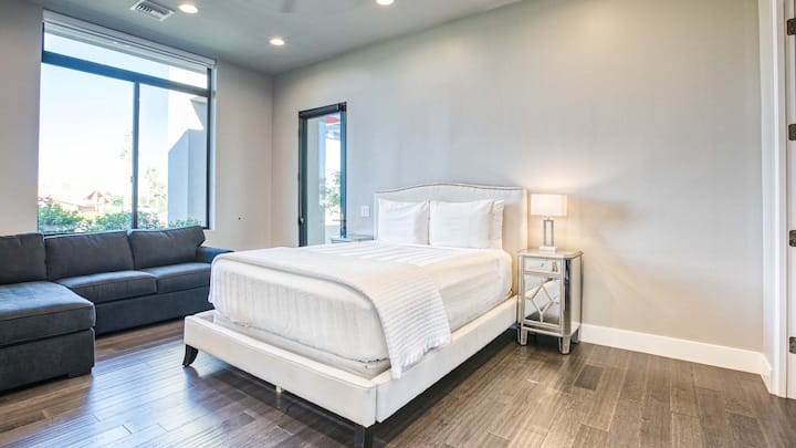 Moderno dormitorio con cama tamaño queen, baño privado, sofá cama gris, grandes ventanales y cálida iluminación empotrada.