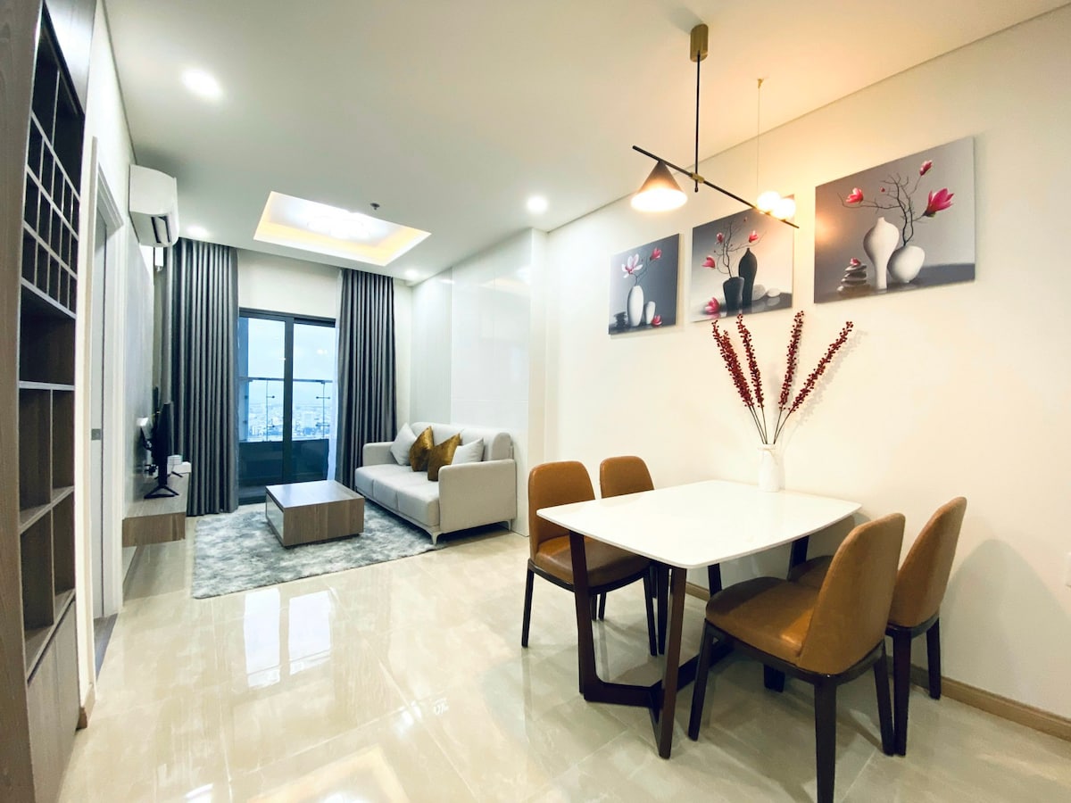 A modern living area features a light-toned sofa paired with a rectangular coffee table. A dining table with four brown chairs occupies the space, decorated with a vase of dried flowers. Two artworks are displayed on the wall, adding a subtle enhancement to the overall decor.