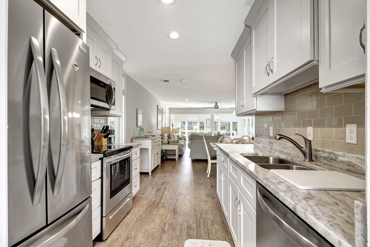 A modern kitchen features stainless steel appliances, including a refrigerator, oven, and microwave. Soft gray cabinetry complements the light-colored countertops. A clear view of the open living area is visible, with natural light streaming through windows, and a cozy seating arrangement in the background.