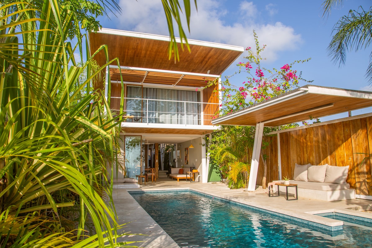 A modern tropical house is surrounded by lush greenery, featuring a private pool in the foreground. The two-story structure includes expansive glass doors that open to the living area, and a shaded seating area with neutral-colored cushions sits under a wooden overhang.