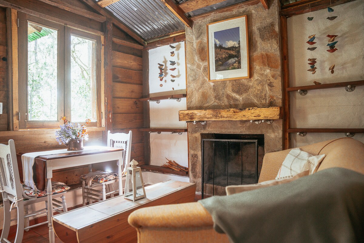 An intimate seating area is showcased with a comfortable couch beside a rustic stone fireplace. A small wooden table with two chairs is positioned near the window, which allows natural light to filter through, illuminating the room's natural materials and decorative elements.