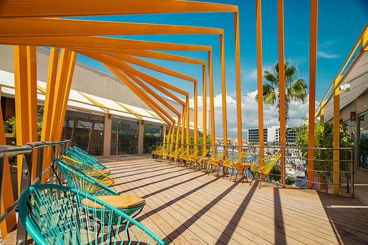 A vibrant outdoor terrace features a series of striking yellow beams creating a sense of depth. Colorful lounge chairs in turquoise are arranged along the wooden deck, complemented by natural light streaming through, highlighting the palm trees in the background and providing a relaxed seating area.