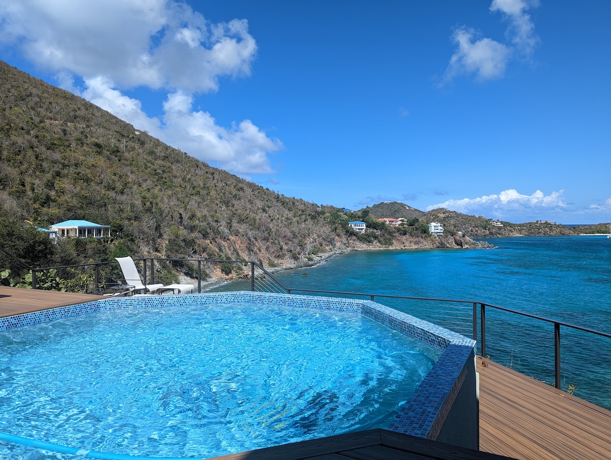 An octagonal pool with a sparkling blue surface is located on a wooden deck overlooking a vibrant bay. The surrounding landscape features lush hills and scenic coastlines, with clear skies and a few white clouds enhancing the view.