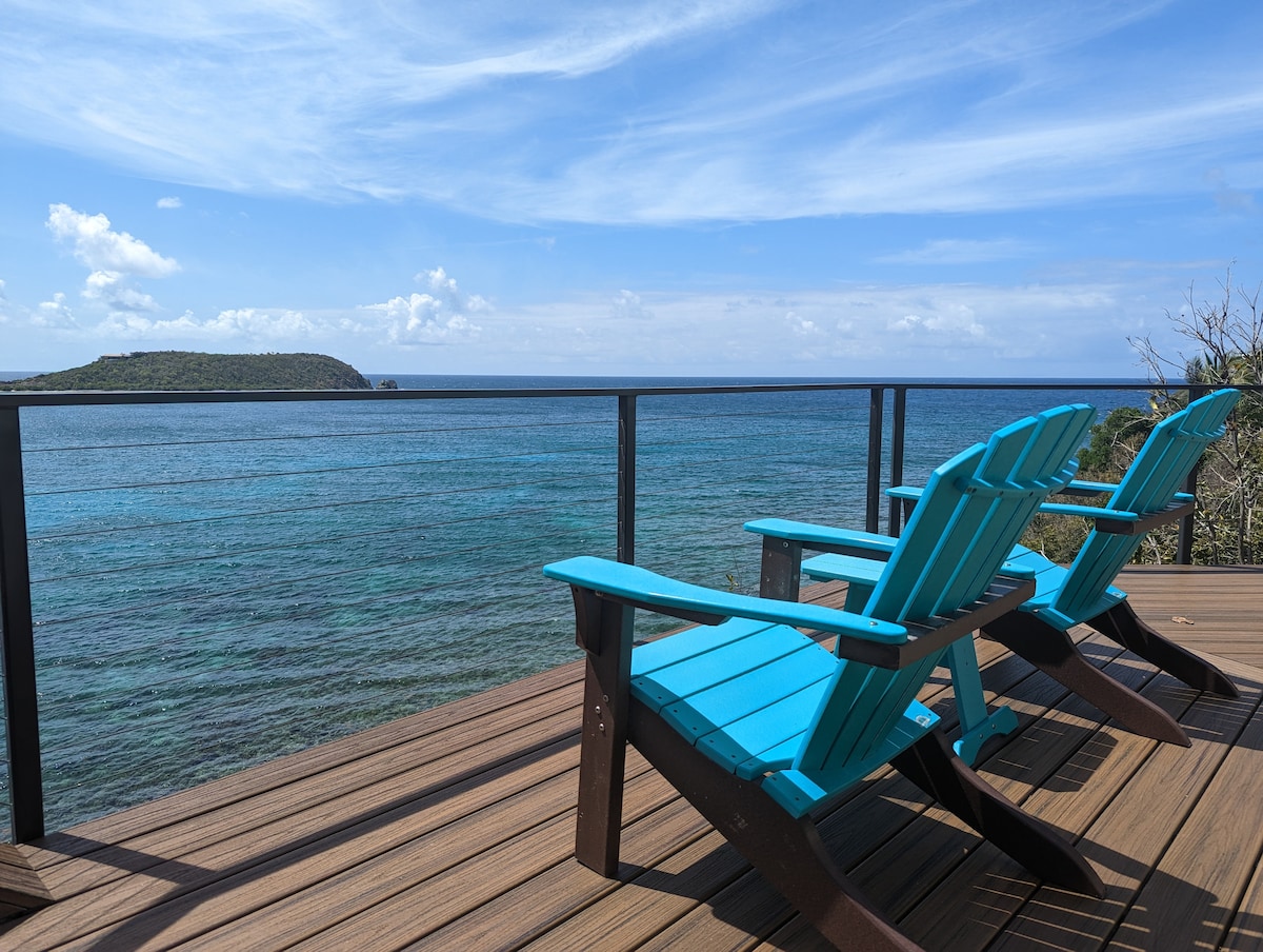 Two vibrant turquoise Adirondack chairs are positioned on a wooden deck, overlooking calm azure waters. The view extends toward distant green hills under a partly cloudy sky, creating a serene coastal atmosphere.