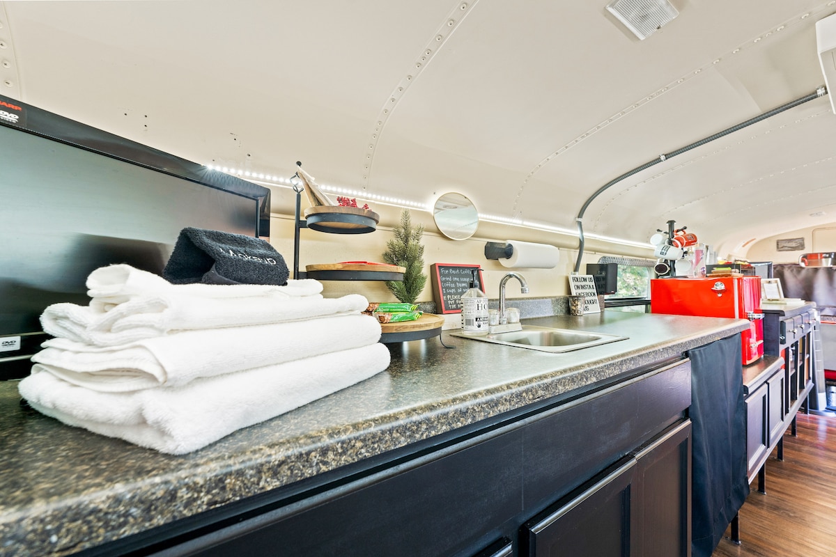 The kitchenette features a well-organized surface with neatly folded white towels, a sink, and a compact coffee maker. Nearby, a small decorative plant and various kitchen items are displayed, providing essential functionality in the cozy bus interior.