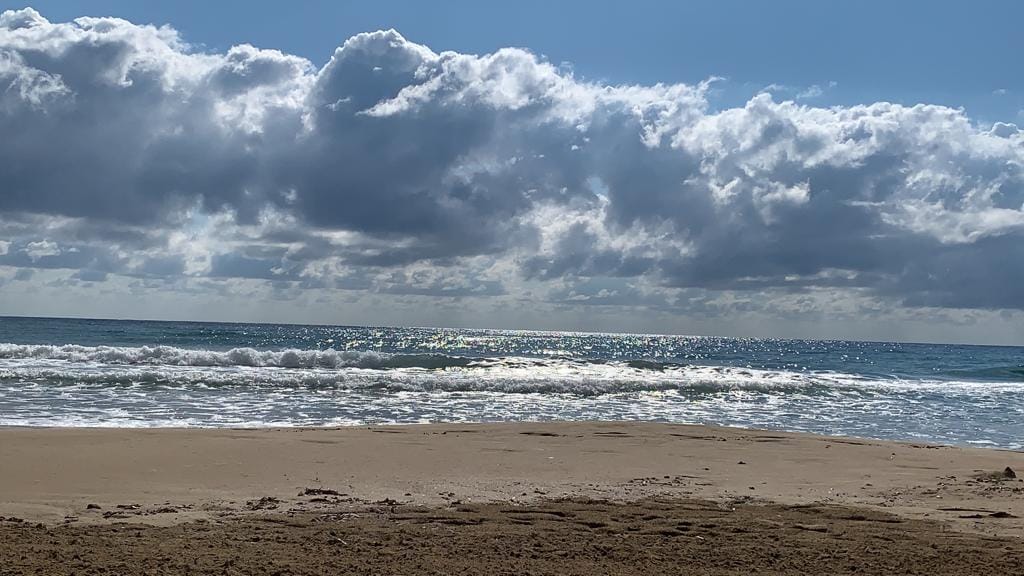 A wide expanse of sandy beach is visible, meeting the tranquil blue waters of the sea. The sky is filled with scattered clouds, allowing sunlight to shimmer on the surface of the waves, creating a serene coastal atmosphere.