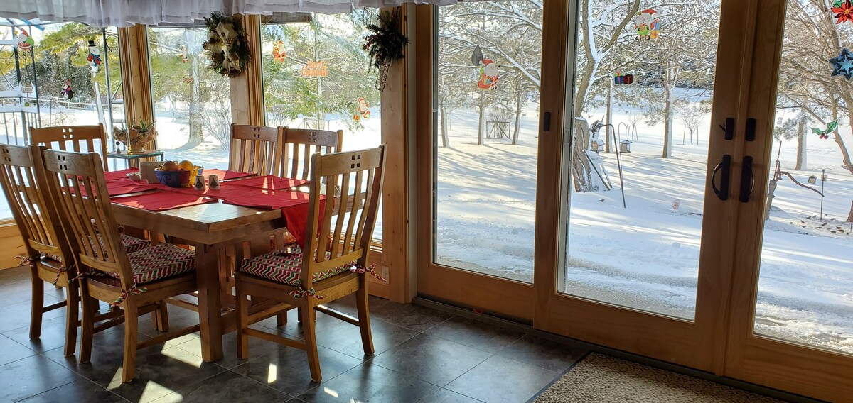 A dining area features a wooden table surrounded by chairs, set with a red tablecloth and decorative items. Large glass doors provide views of a snowy outdoors, with a serene landscape visible through the transparent panels, enhancing the natural light within the space.