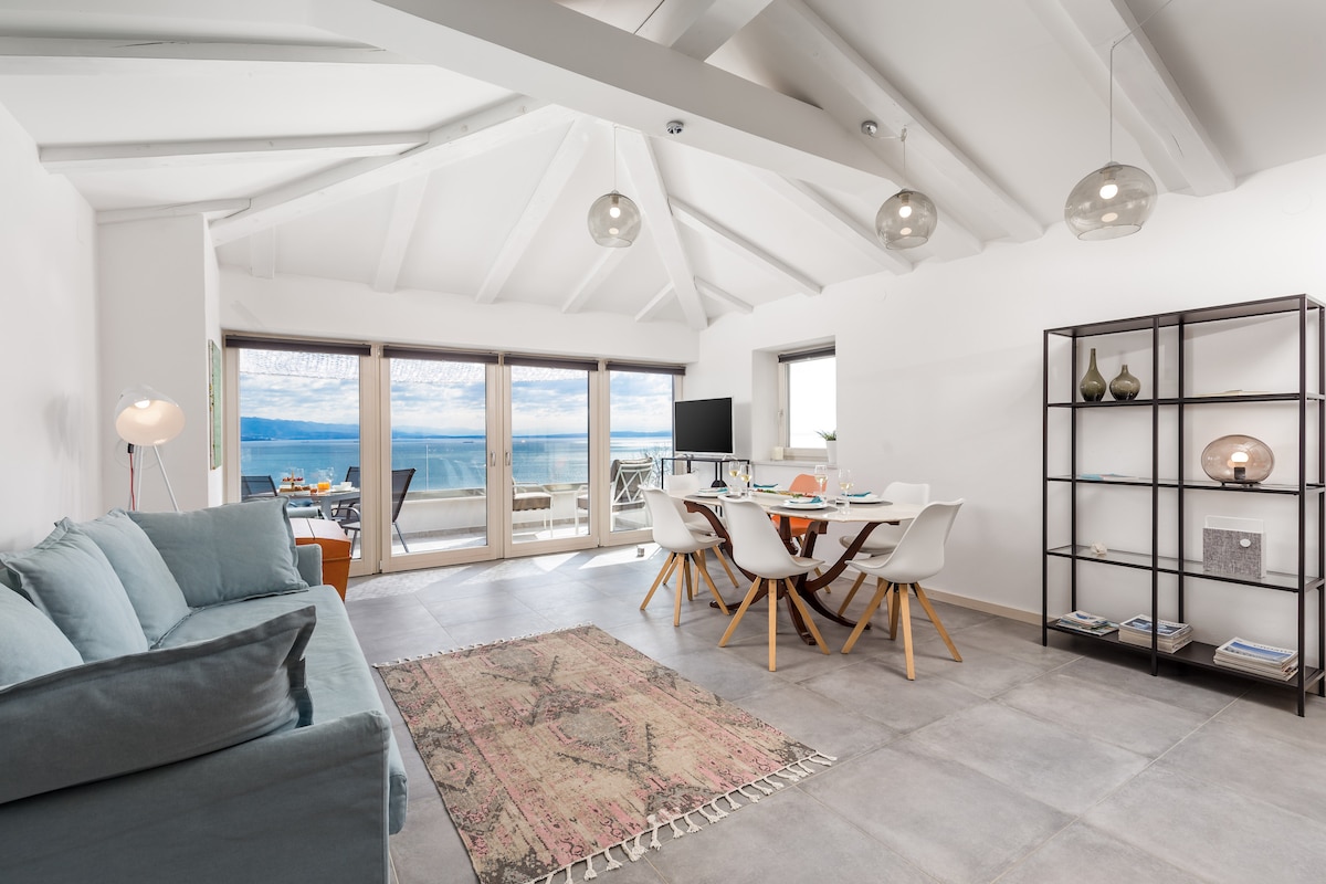 A loft-style living area is illuminated by natural light streaming through large glass doors, offering views of the sea. Comfortable seating is arranged around a table that accommodates multiple guests. Modern decor includes a patterned rug and minimalist shelving, enhancing the spacious feel.