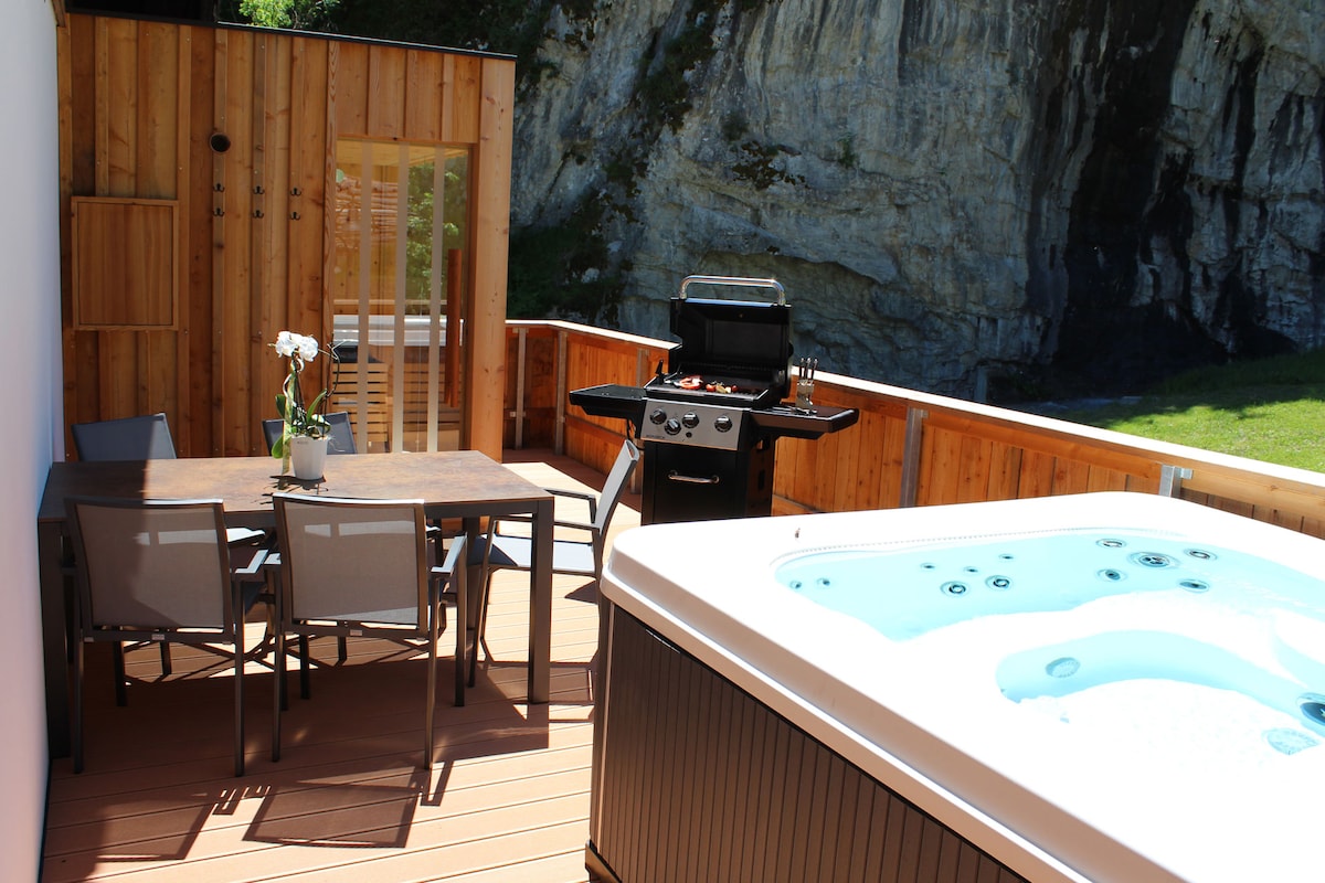 An outdoor terrace features a spacious hot tub, a modern barbecue grill, and a wooden dining table surrounded by four chairs. The setting is complemented by natural stone and greenery in the background, creating a relaxing outdoor environment.