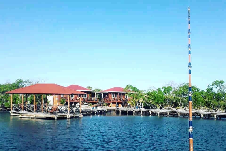 Two private open-style cabanas are visible on a lush, remote island, featuring red roofs and wood-paneled construction. Surrounding them, greenery enhances the serene setting. The calm waters of the Belize Barrier Reef reflect the clear blue sky, providing a tranquil backdrop to the island retreat.