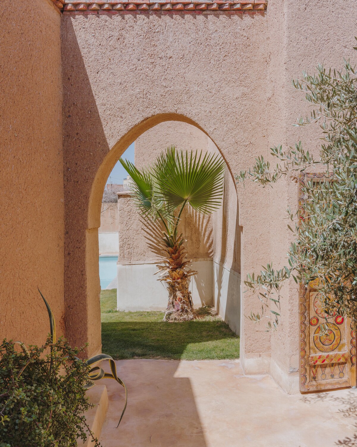 A passageway framed by traditional adobe walls guides the viewer towards a picturesque oasis. A palm tree stands prominently in the foreground, while a glimpse of the pool can be seen beyond, surrounded by lush greenery.