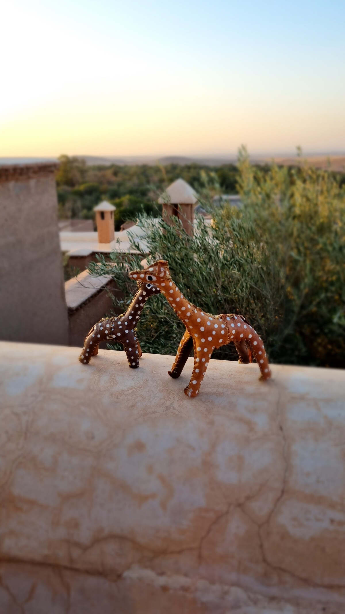 Two decorative giraffe figurines, one brown with white spots and the other in a darker shade, are positioned on a ledge overlooking greenery. The background features a soft sunset glow, enhancing the serene landscape.