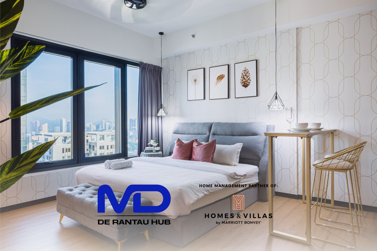 A spacious studio suite is featured, showcasing a king bed with a grey upholstered headboard. Large windows allow natural light to illuminate the area, complemented by minimalistic decor. A cozy seating area with a small ottoman and bedside lighting adds to the functional space.