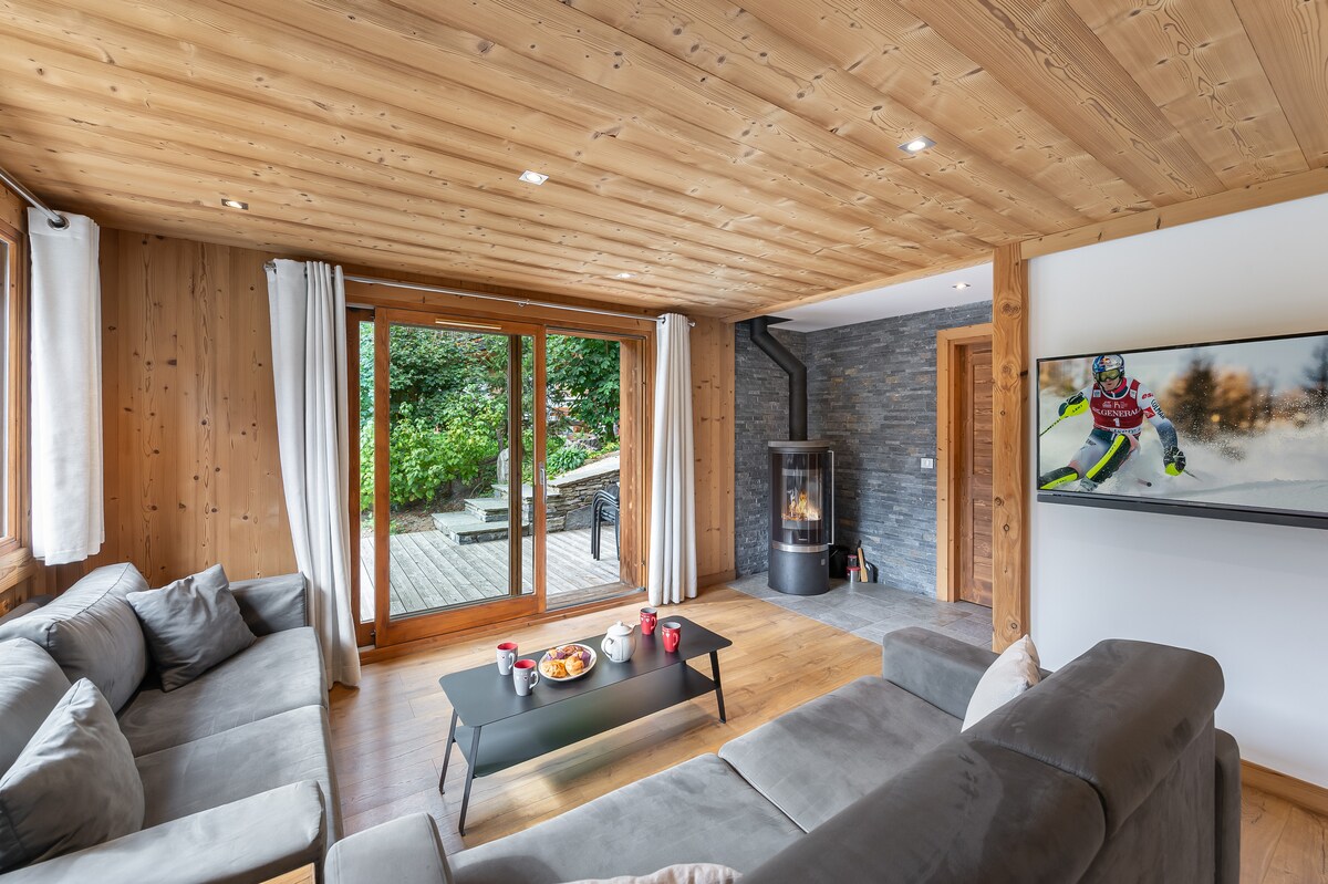A cozy living area showcases a large, comfortable sectional sofa, complemented by a black coffee table. Natural light floods in through expansive glass doors that open to an outdoor terrace. A wood-burning stove and wall-mounted TV add to the inviting atmosphere of the space.