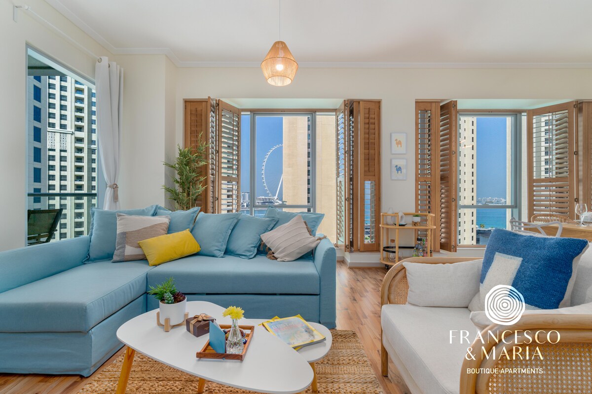 A bright and airy living area features a large sectional sofa in soft blue hues and a light-colored armchair. Floor-to-ceiling windows with wooden shutters allow ample natural light and showcase views of Ain Dubai. A stylish coffee table holds decorative items and a small plant.