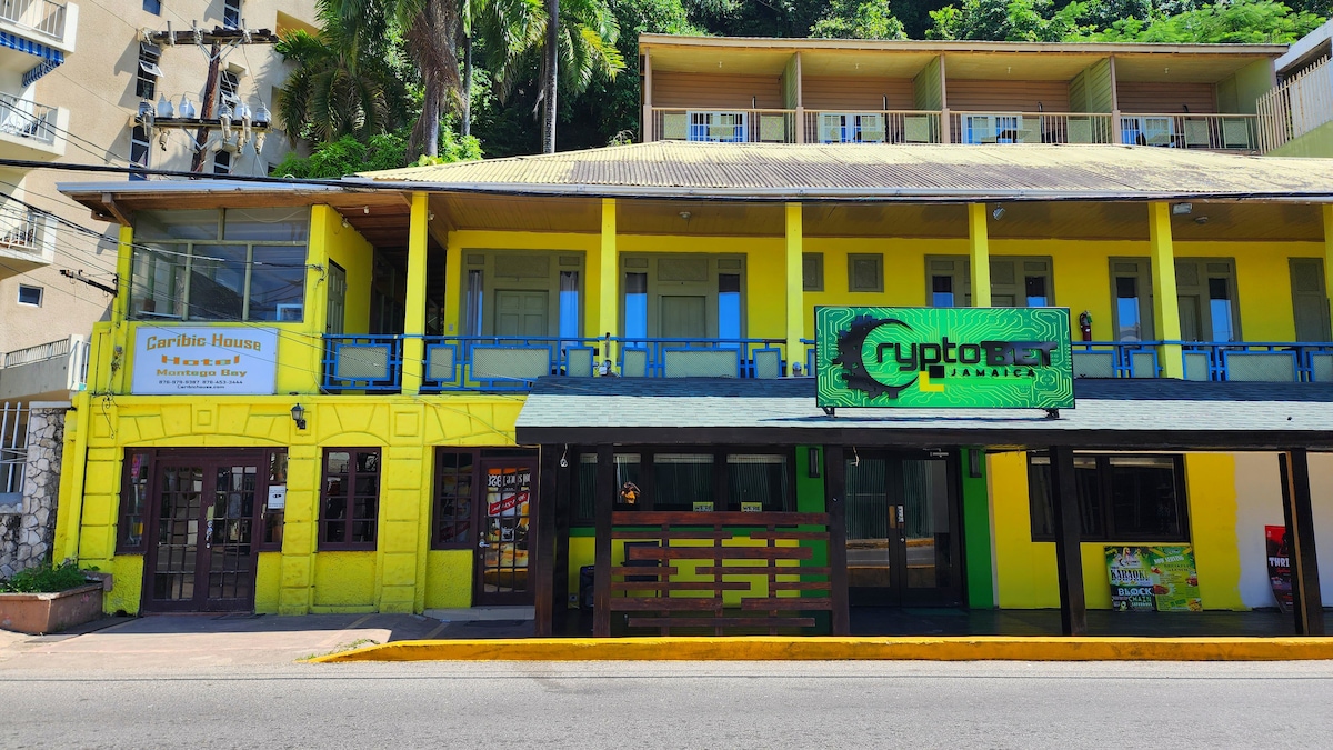 A vibrant multi-story building features bright yellow exteriors and a spacious terrace with a dark wood railing. The ground level includes a storefront and an entrance to the hotel, while the second level displays a series of blue-framed windows and a sign for a local business.