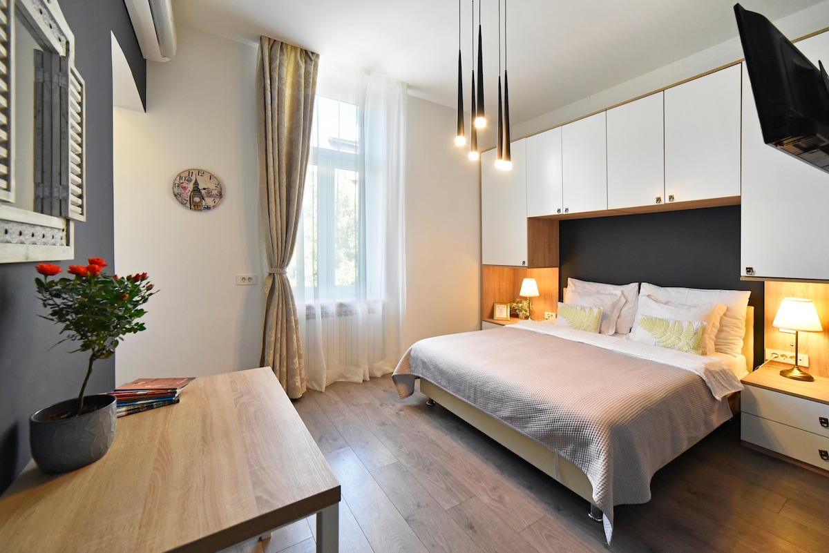 A modern room features a spacious bed with light bedding, against a dark accent wall. Two bedside lamps provide gentle illumination. A wooden desk is positioned nearby, complemented by a small plant. Floor-to-ceiling curtains frame the window, allowing natural light to enhance the space.