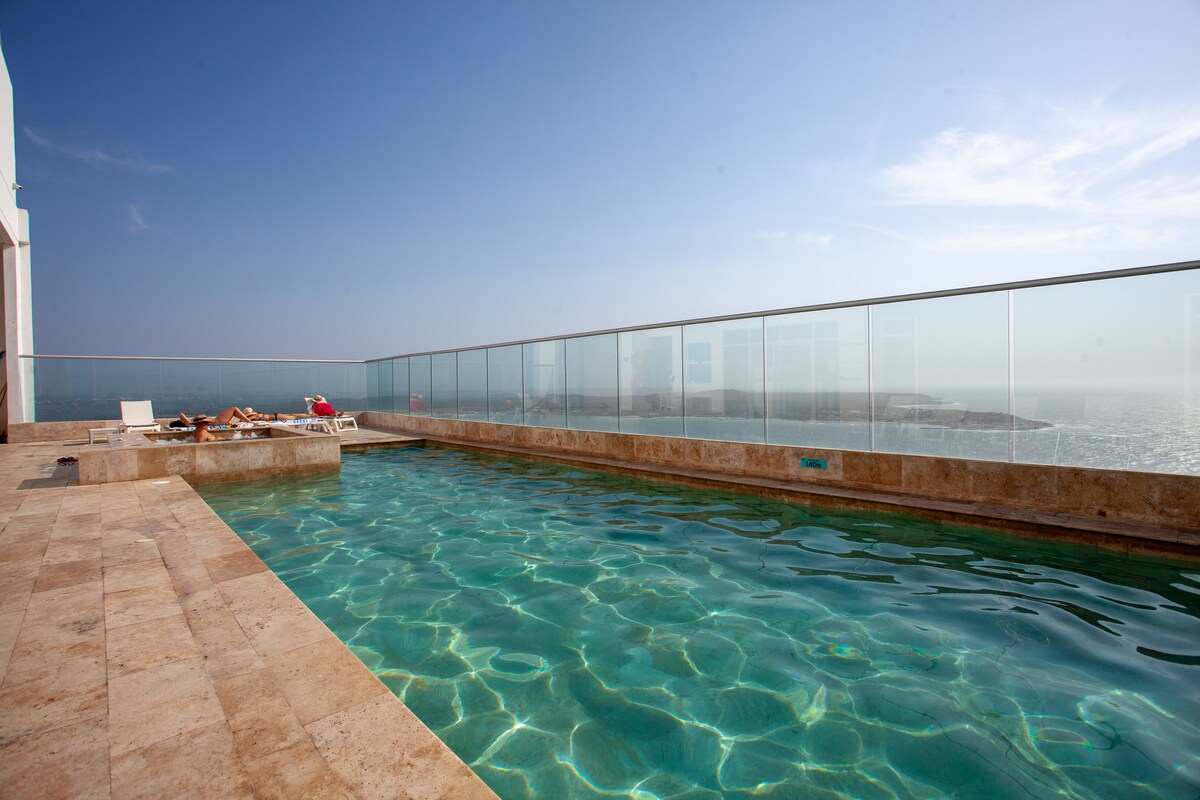 An infinity pool is visible, bordered by glass, offering unobstructed views of the sea. The pool’s water is clear, reflecting the sky. Surrounding the pool area are stone tiles, with several lounge chairs positioned nearby for relaxation.