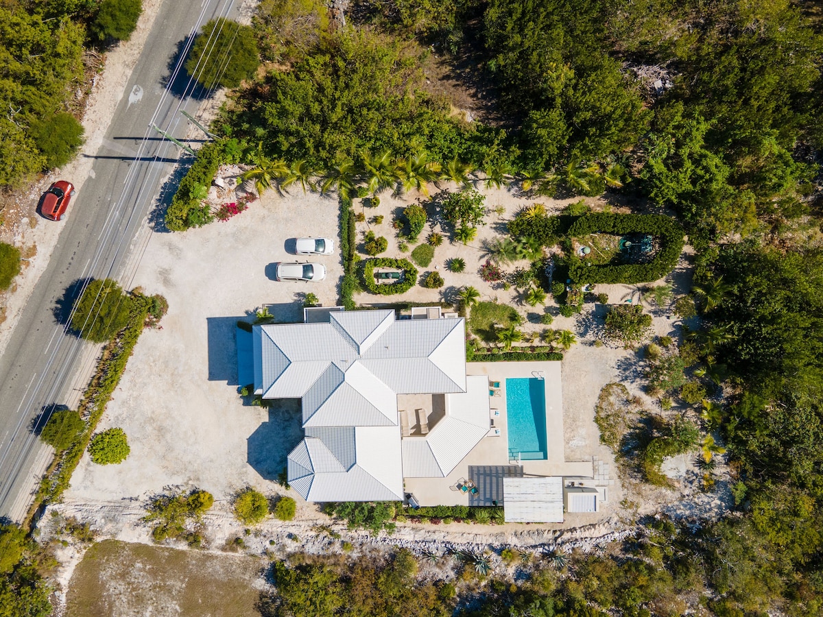 An aerial view captures the villa situated amidst lush greenery, with a well-maintained garden surrounding the spacious property. A private pool is visible in the rear, alongside a patio area for outdoor relaxation. The nearby road provides easy access to this serene retreat.