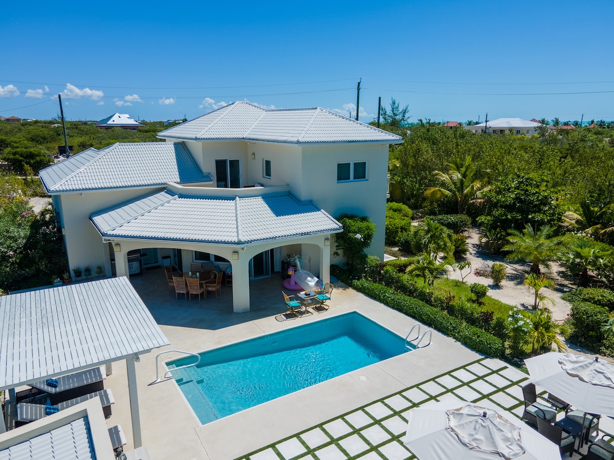 An aerial view of a modern villa showcases a rectangular pool surrounded by a smooth patio. A shaded outdoor seating area can be seen adjacent to the pool. Lush greenery and well-manicured landscaping create a serene setting for relaxation.