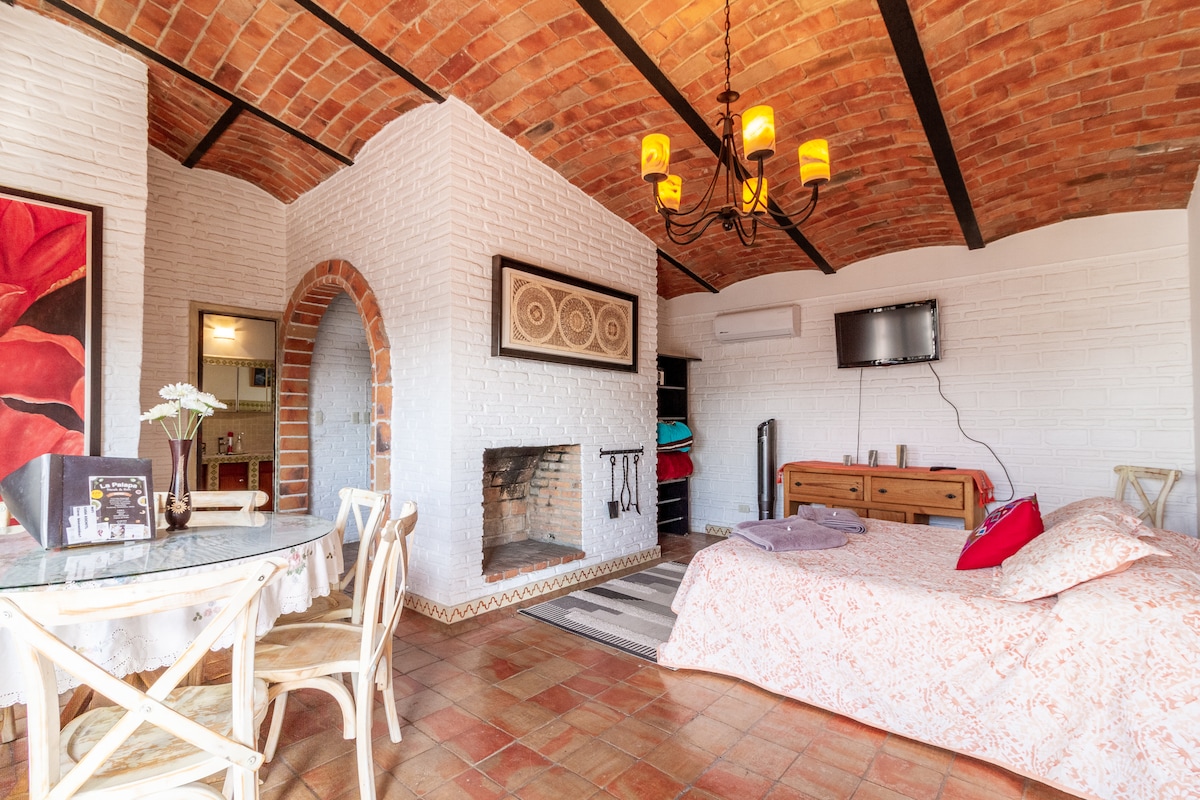 A cozy room features a spacious bed with a light patterned coverlet, surrounded by warm brick walls and accented by exposed wooden beams. A small dining area includes a glass table and rustic chairs. A decorative fireplace provides warmth, and a wall-mounted TV is visible.