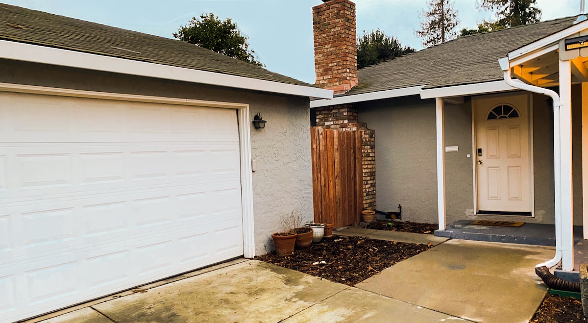The exterior of the duplex presents a single-car garage and an inviting entryway. A welcoming front door is flanked by a small porch. Potted plants and a fenced area add to the outdoor space, with a charming chimney visible on the siding.