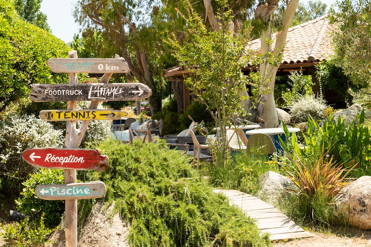 Multiple directional signs are positioned amidst lush greenery, indicating various facilities such as tennis, reception, and the pool. A charming wooden footpath leads through the garden, surrounded by vibrant plants and trees that create a serene outdoor environment.