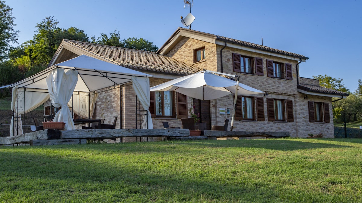 A two-story villa is showcased amidst a well-maintained green lawn. The exterior features rustic brick walls and brown shutters. Adjacent to the house, a covered outdoor seating area is set up with white canopies, providing shade on sunny days.