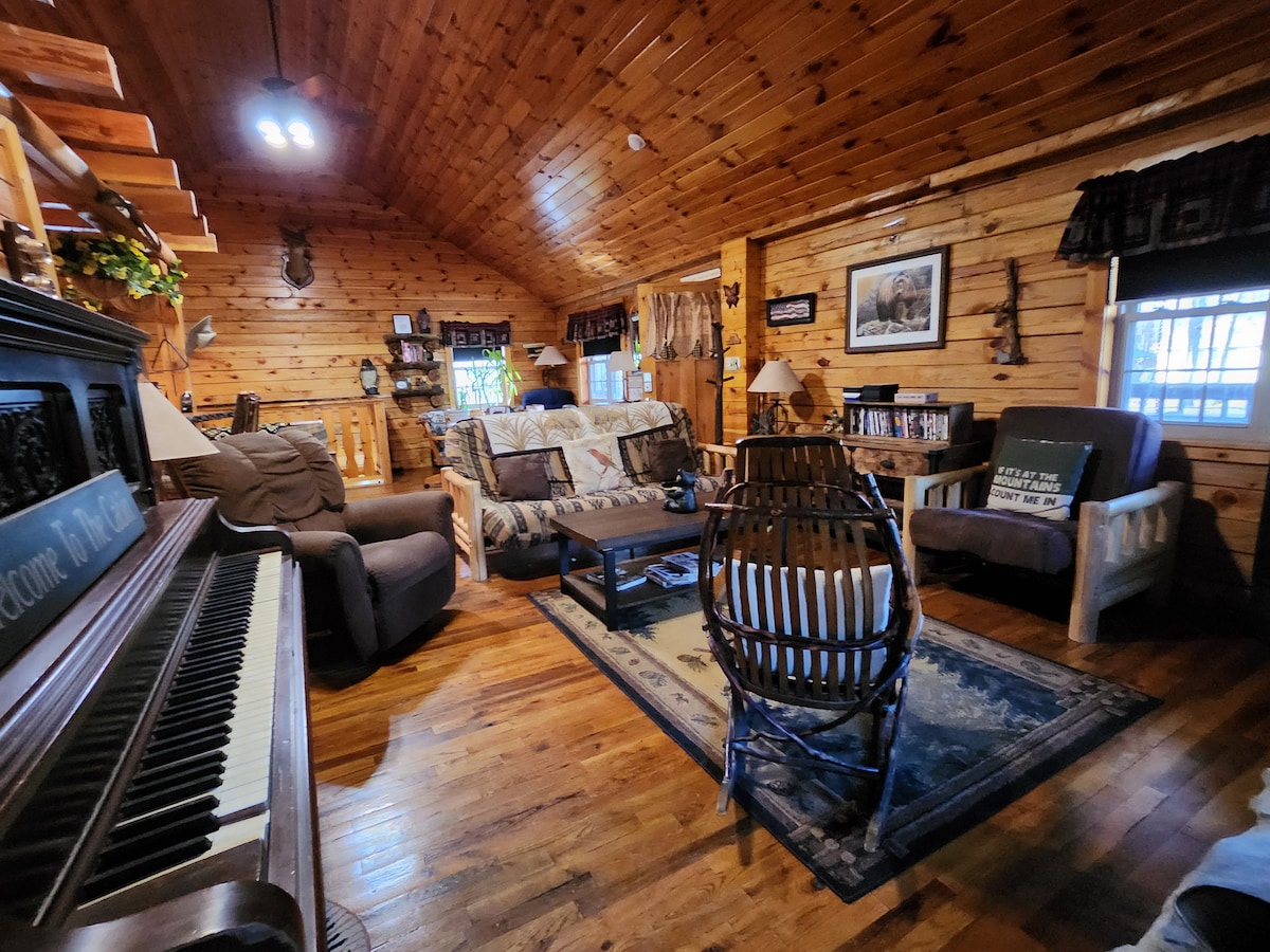 A warm and inviting living area features wooden walls and a high ceiling, enhancing a rustic charm. Comfortable seating options include a couch, armchairs, and a wicker chair. A piano rests against one wall, alongside decorative items and framed pictures, creating a cozy atmosphere.