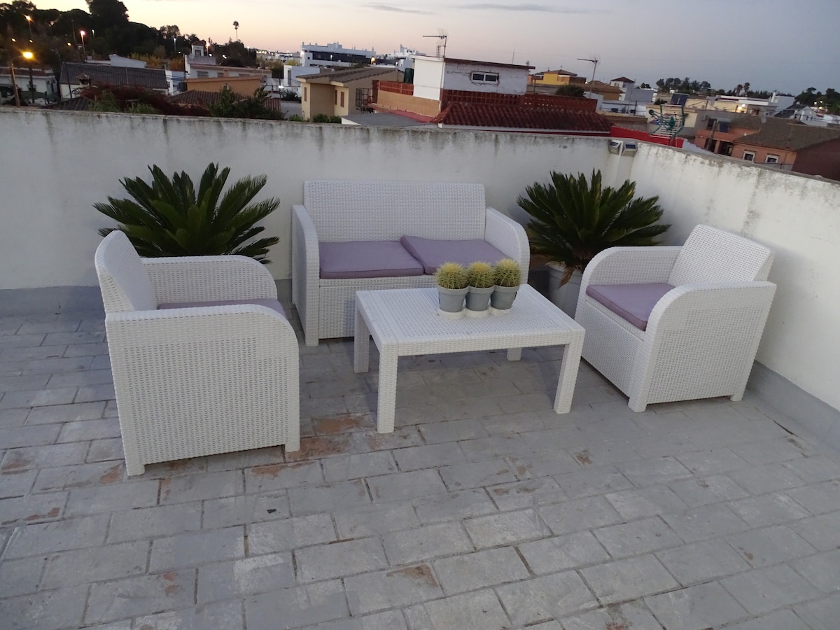A terrace area features a seating arrangement consisting of a white outdoor sofa set, accompanied by matching armchairs. A coffee table is positioned in the center, with small potted plants placed on it. The surrounding view includes rooftops and distant hills under a soft evening sky.