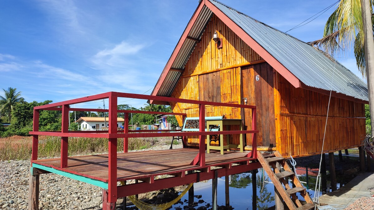 A traditional wooden hut features an elevated deck with a red railing, overlooking a calm waterway. The structure's sloped roof is made of metal, and natural wooden panels create a warm appearance. Lush greenery is visible in the background, enhancing the rural setting.