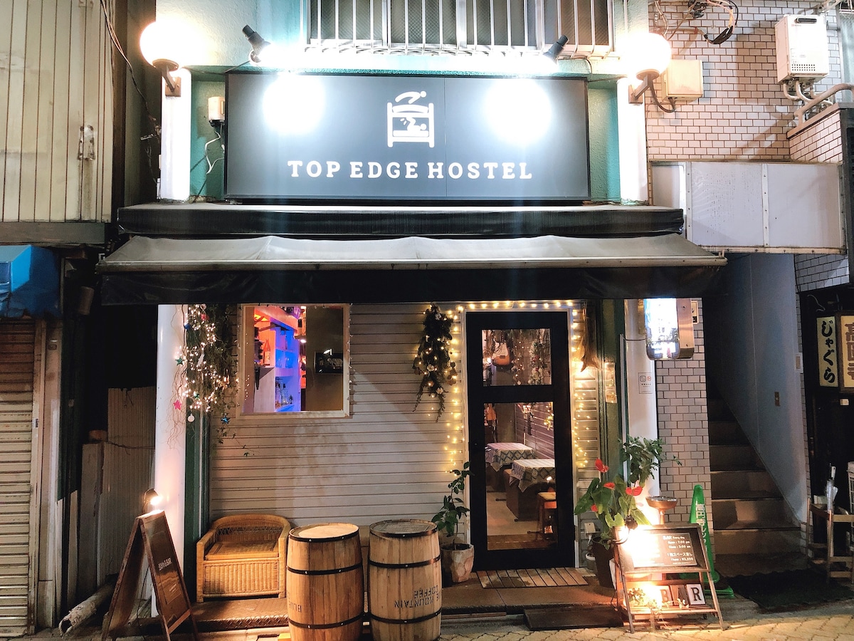 The exterior of the Top Edge Hostel is shown, featuring a welcoming entrance with a large sign overhead. Wooden barrels are placed at the door, and soft lighting decorates the front, creating an inviting atmosphere. The urban setting includes neighboring buildings and street elements.