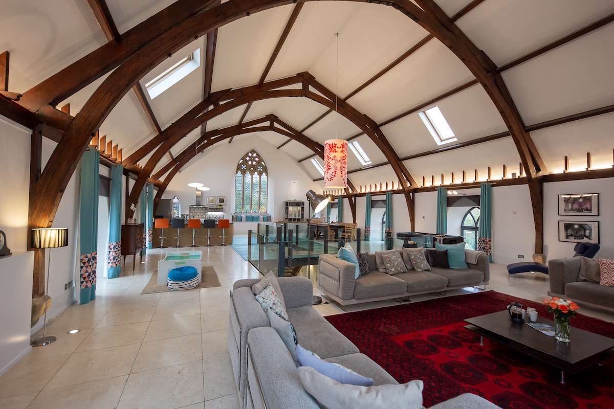 An open plan living area features a high vaulted ceiling with exposed wooden beams. Elegant sofas are arranged around a coffee table, while a vibrant red area rug lies centrally. Natural light flows through large windows, highlighting the contemporary kitchen area in the background.