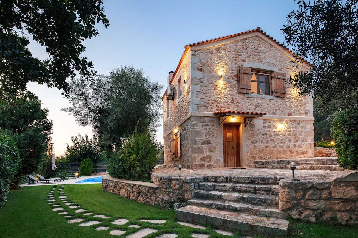 A stone-built villa is depicted, showcasing a rustic exterior with wooden shutters and a welcoming entrance. Pathways lead to the front door, surrounded by landscaped gardens. A private swimming pool is visible in the background, framed by lush greenery, creating an inviting atmosphere.