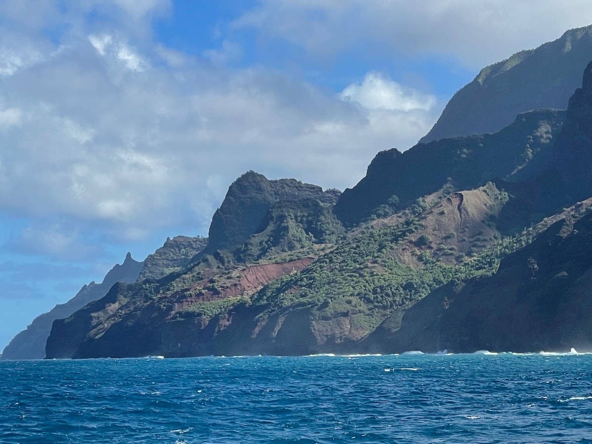 A scenic view captures rugged coastal cliffs rising dramatically from the ocean. Towering green hillsides are interspersed with rocky formations under a partly cloudy blue sky. Waves crash gently against the shore, creating a serene atmosphere.
