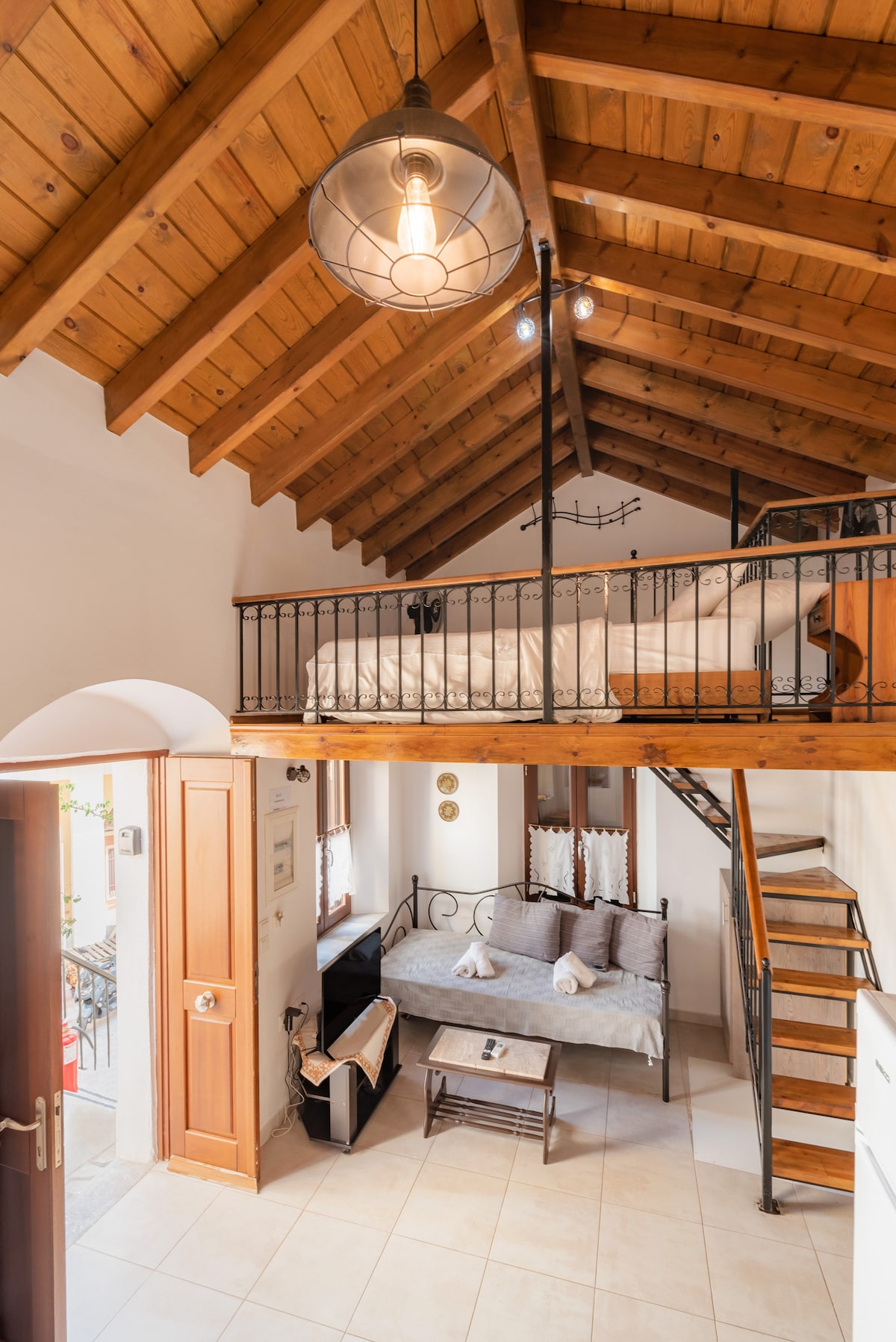 The interior features a high wooden ceiling with exposed beams. A cozy sleeping area is positioned on a loft above, while a sitting space with a couch and coffee table is located below. Natural light fills the room, creating a warm, inviting ambiance.