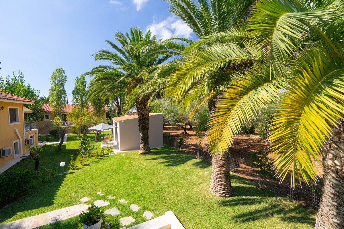 A lush garden area is displayed, featuring palm trees and well-maintained grass. A shaded gazebo is positioned within the garden. The cheerful greenery creates a serene environment, with clear blue skies above, enhancing the outdoor space of the villas.
