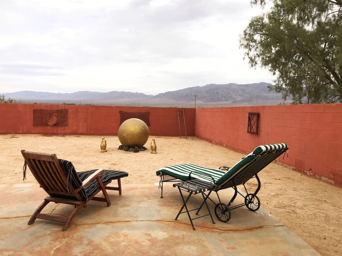 A serene outdoor area features two lounging chairs positioned on a sun-drenched patio. A large golden orb is centered within a fenced space, complemented by views of distant mountains under a partly cloudy sky.