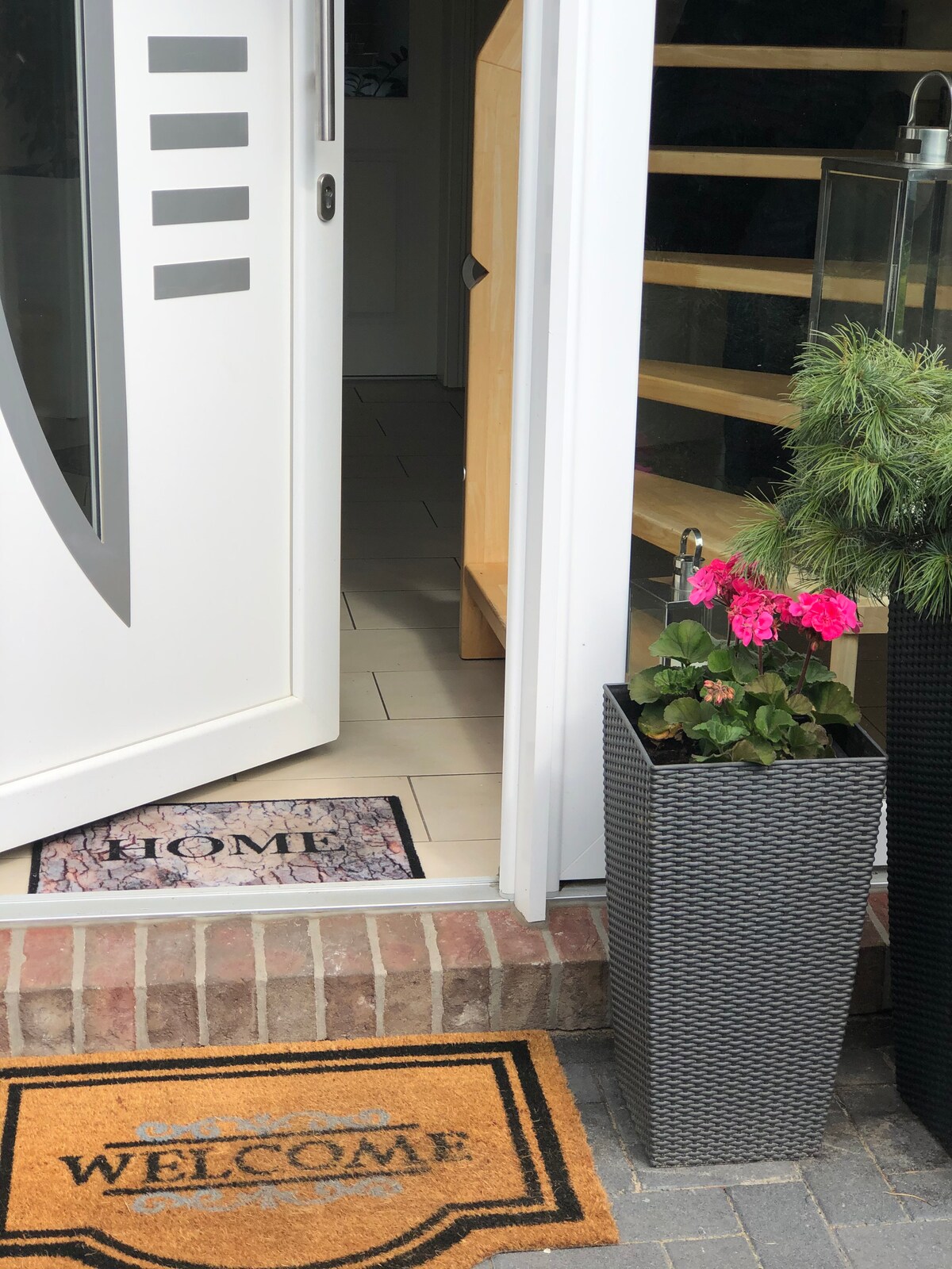 The entrance features a white door open to reveal a light-colored interior. Two welcome mats with distinct messages are placed on the doorstep, alongside a planter with vibrant pink flowers. A decorative lantern adds charm to the outdoor area.