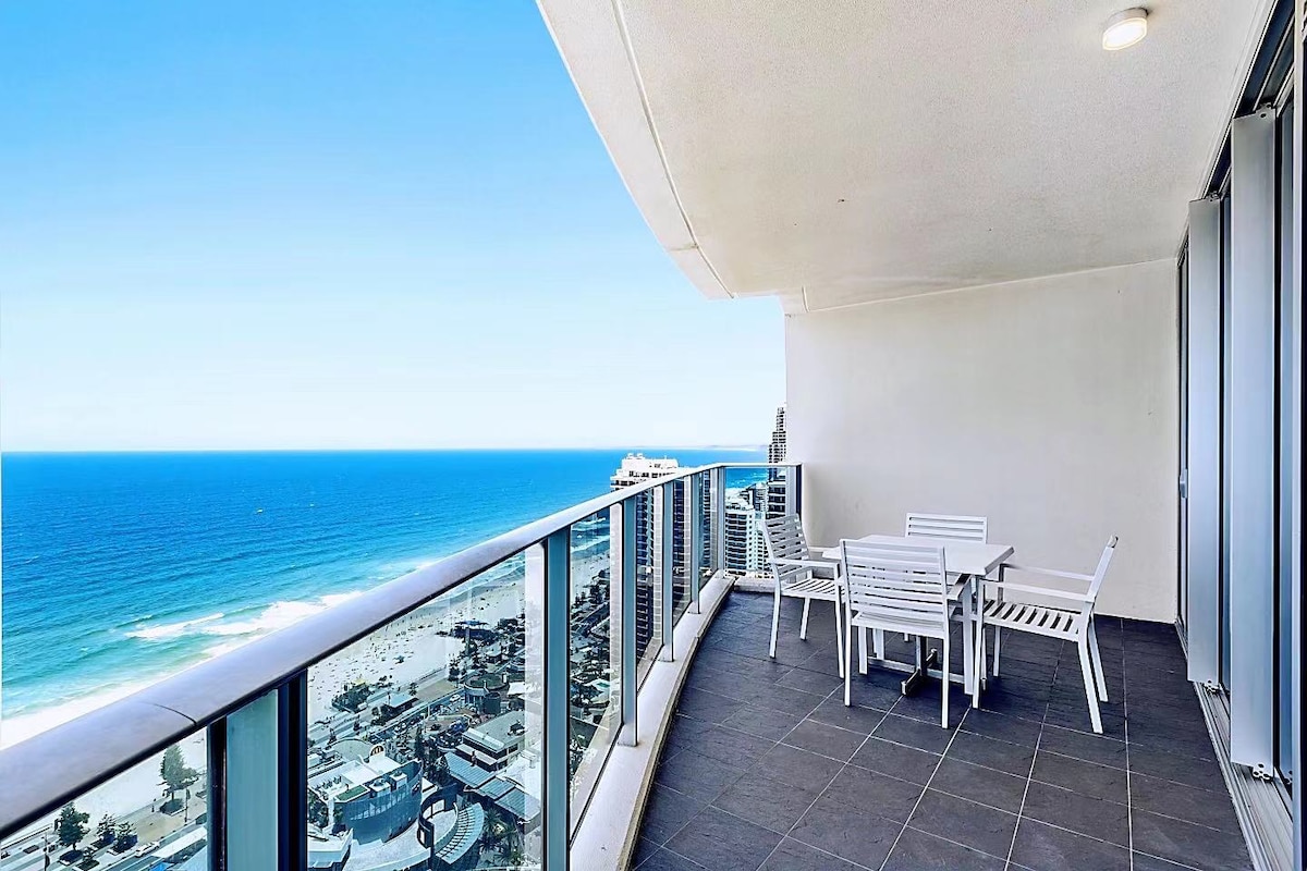 A spacious private balcony features a dining table with several chairs, overlooking a stunning view of the ocean and shoreline. The glass railing provides an unobstructed view, allowing ample natural light to flood the area.