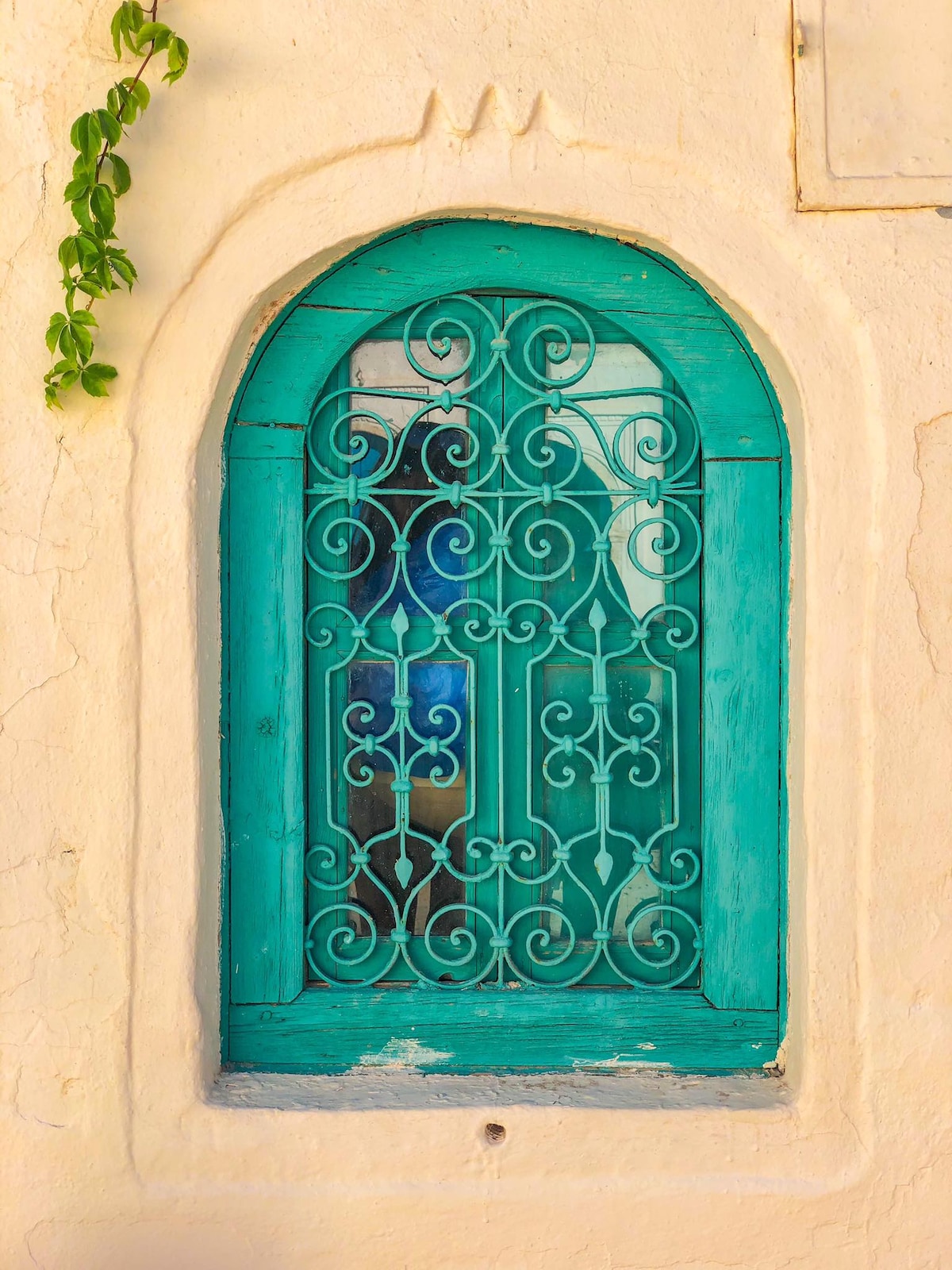 An intricately designed turquoise window with decorative ironwork is set against a textured white wall. Soft light filters through the patterned grill, casting gentle shadows nearby. Ivy softly climbs the side of the window, adding a touch of greenery to the scene.
