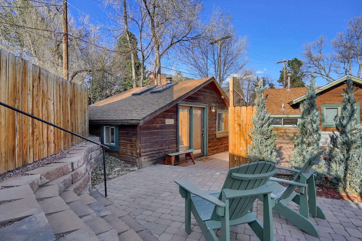 A rustic cottage is positioned within a landscaped area, featuring wooden siding and a welcoming entrance. A paved pathway leads to the cottage, while green chairs offer a seating area. Surrounding trees and a wooden fence create a secluded outdoor space.