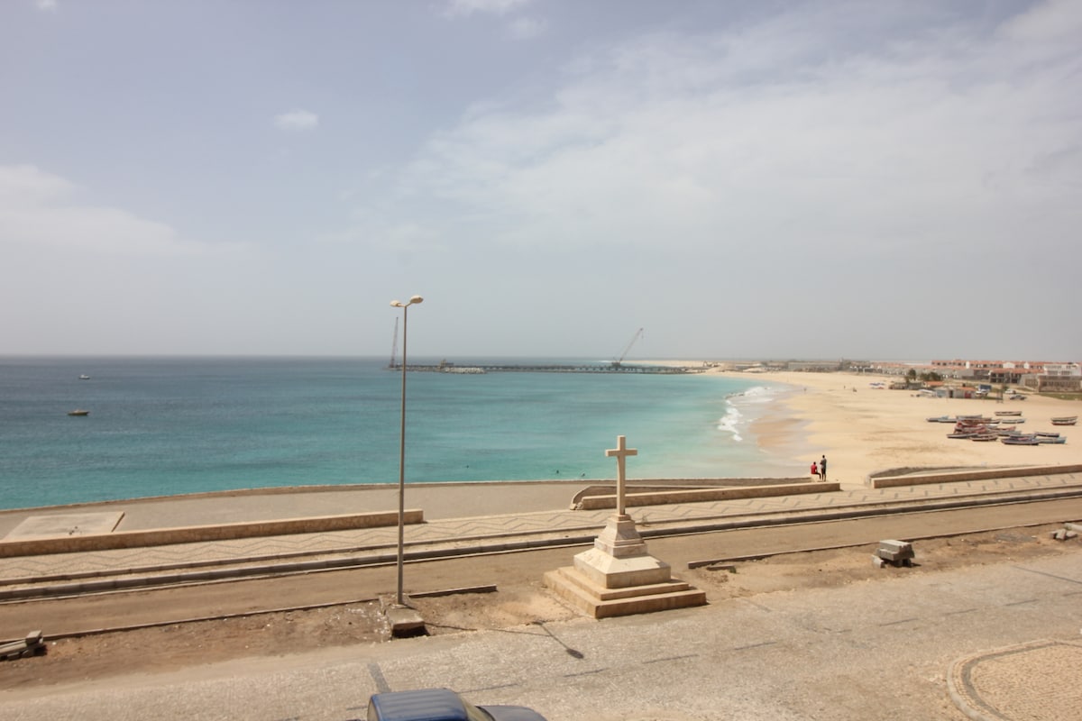 A panoramic view of a sandy beach meets the shimmering blue ocean under a cloudy sky. A pier is visible in the distance, alongside small boats scattered along the shoreline. A cross stands in the foreground, enhancing the tranquil coastal setting.