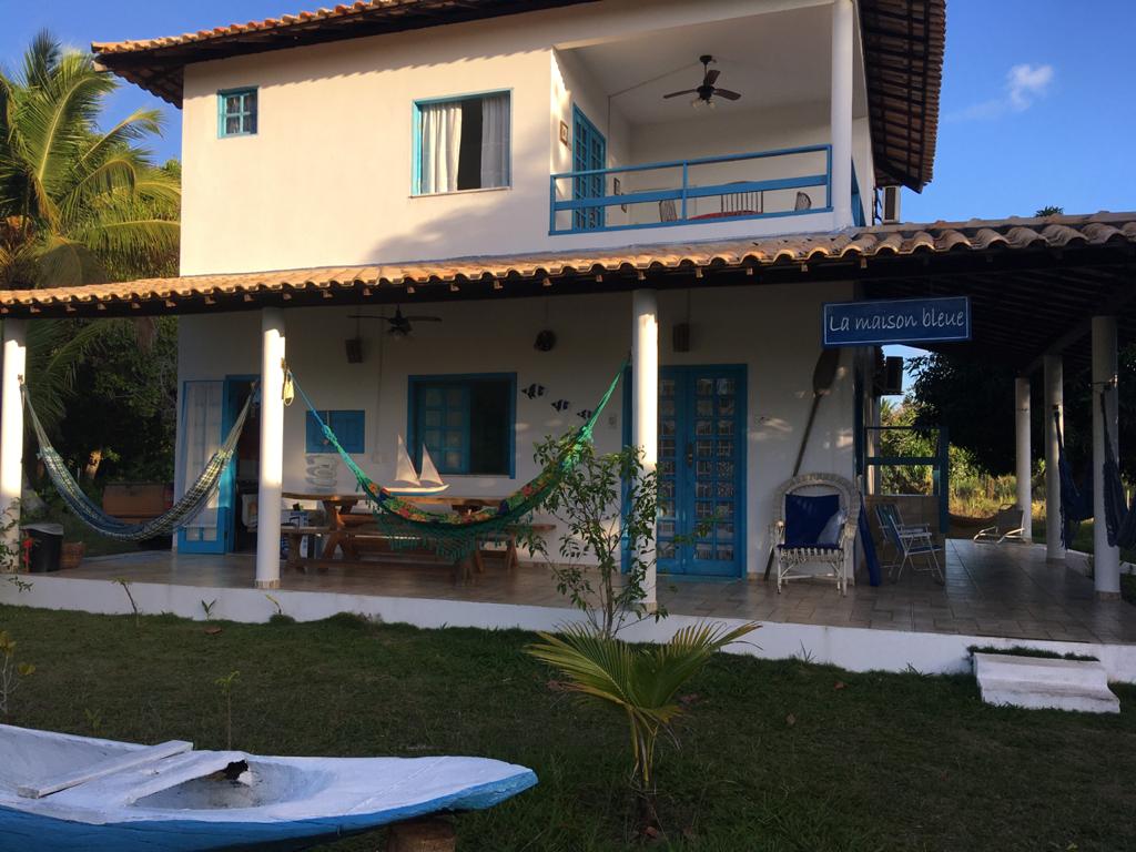 The exterior of the flat features a two-story structure with a white façade and blue window frames. Hammocks are hung on the patio, providing a relaxing space. A sign reading 'La maison bleu' is displayed prominently, surrounded by landscaped greenery.