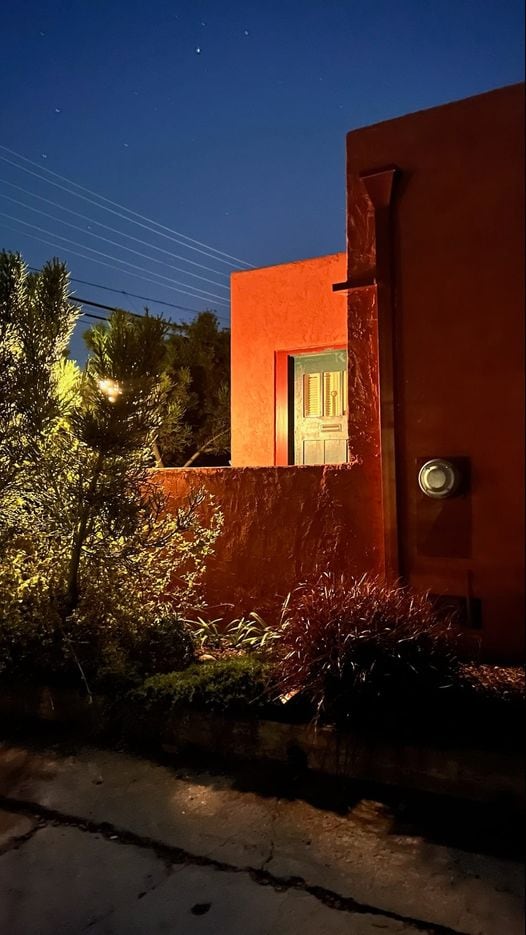 The exterior of the studio is highlighted at night, featuring vibrant red walls and a warm wooden door. Lush greenery surrounds the entrance, including shrubs and ornamental grasses, creating a welcoming ambiance against the softly lit backdrop.