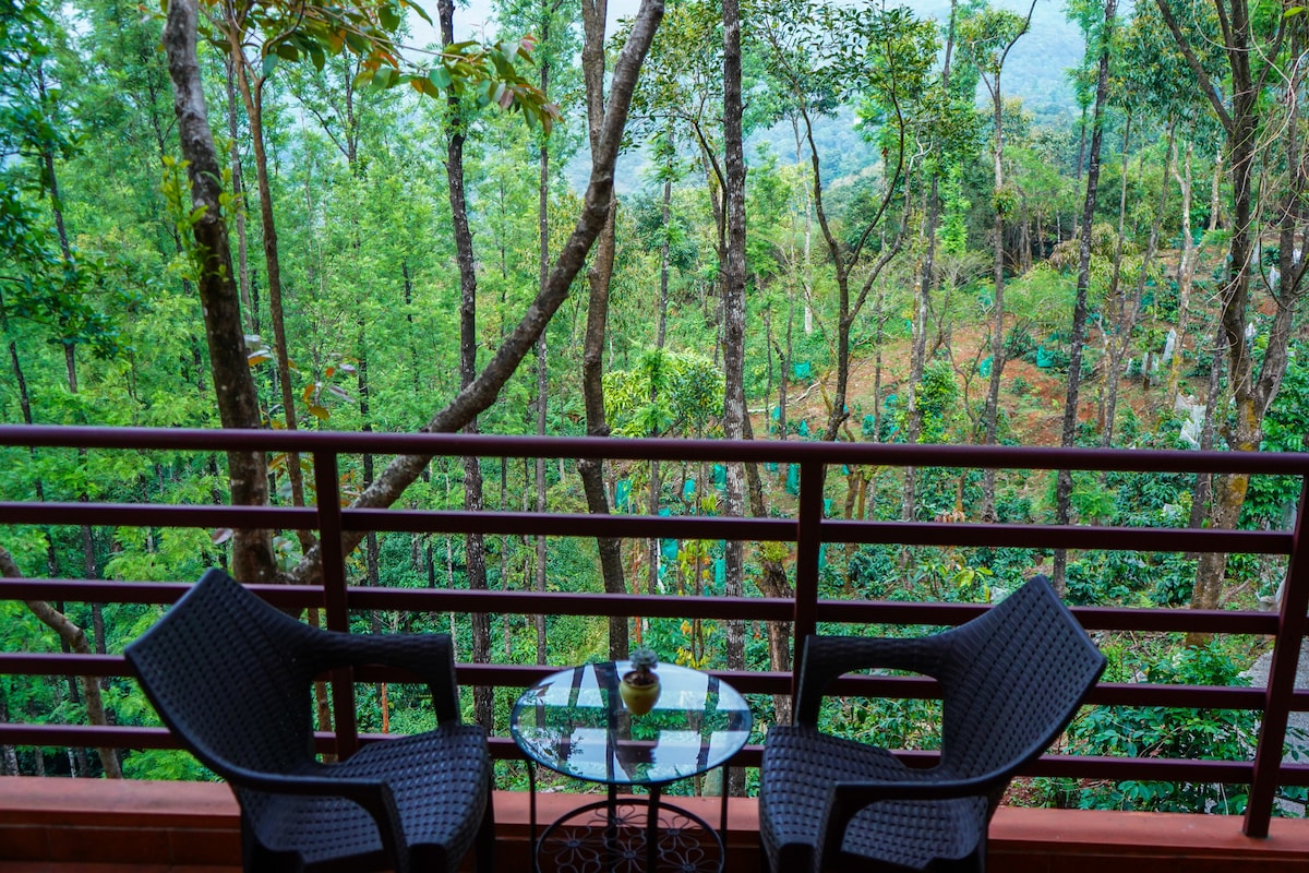 A balcony is seen with two woven chairs and a small glass table, overlooking a lush green landscape filled with trees and vegetation. The natural setting enhances the sense of tranquility and connection to nature.