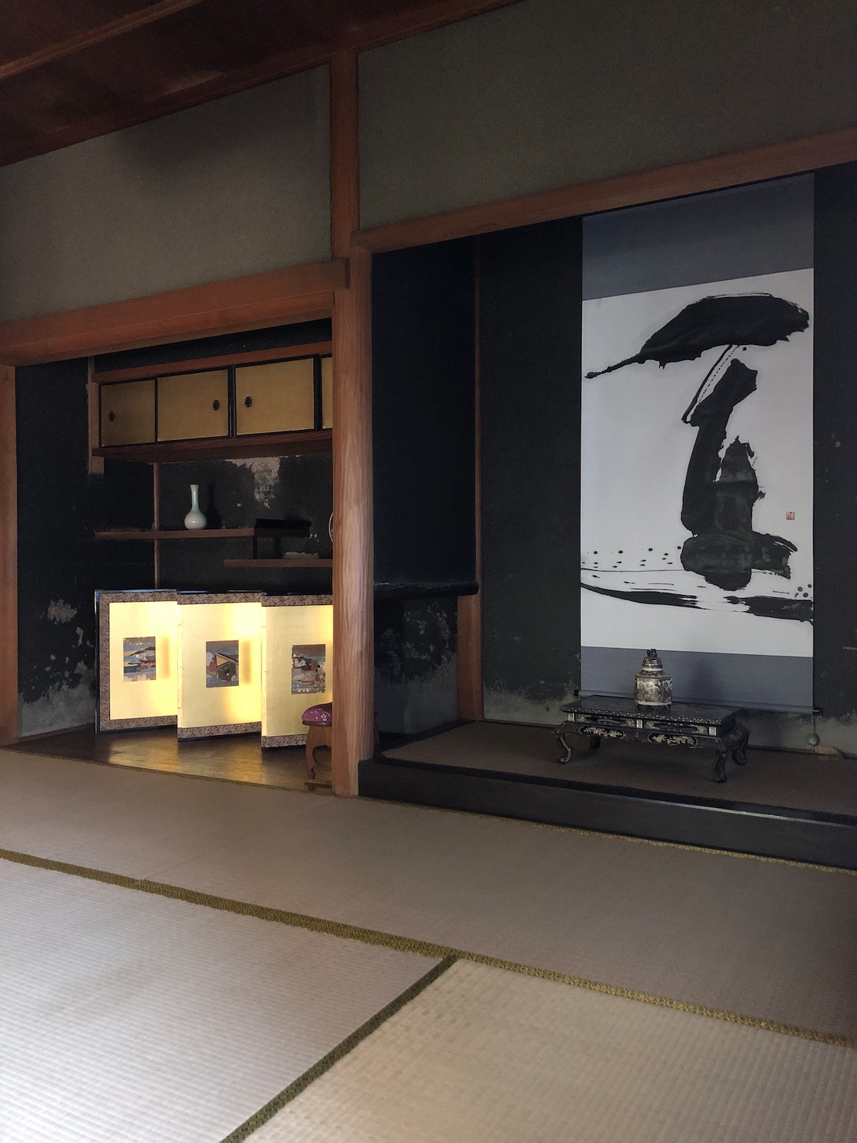 An interior space of a traditional Japanese home is featured, showcasing tatami flooring and wooden architecture. A wall scroll with a brush painting is displayed prominently. A small shelf with decorative items is positioned against the wall, providing a glimpse into the room's cultural aesthetics.