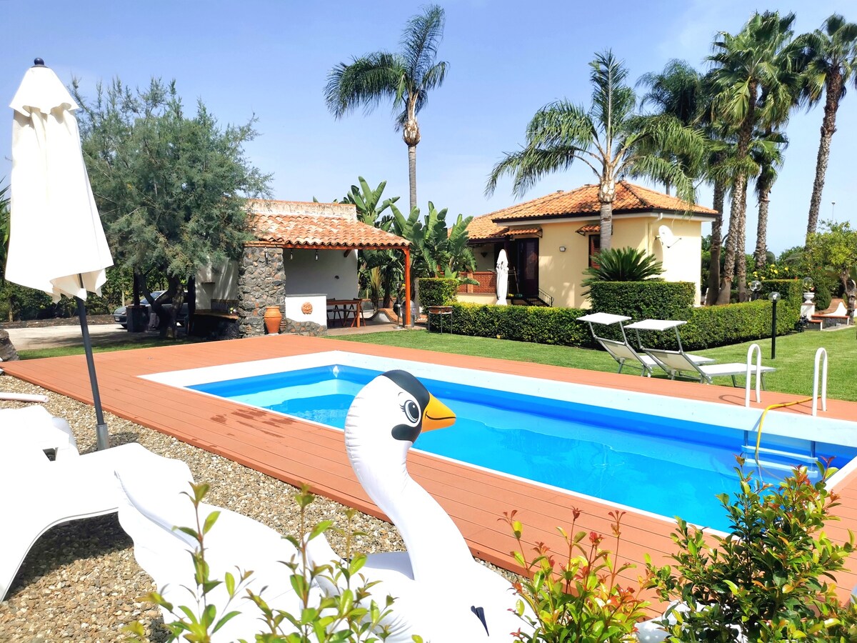 A private swimming pool is flanked by lounge chairs on a wooden deck, with tropical palm trees in the background. A charming stone structure is visible, featuring an outdoor dining area shaded by an umbrella, set amidst lush greenery.