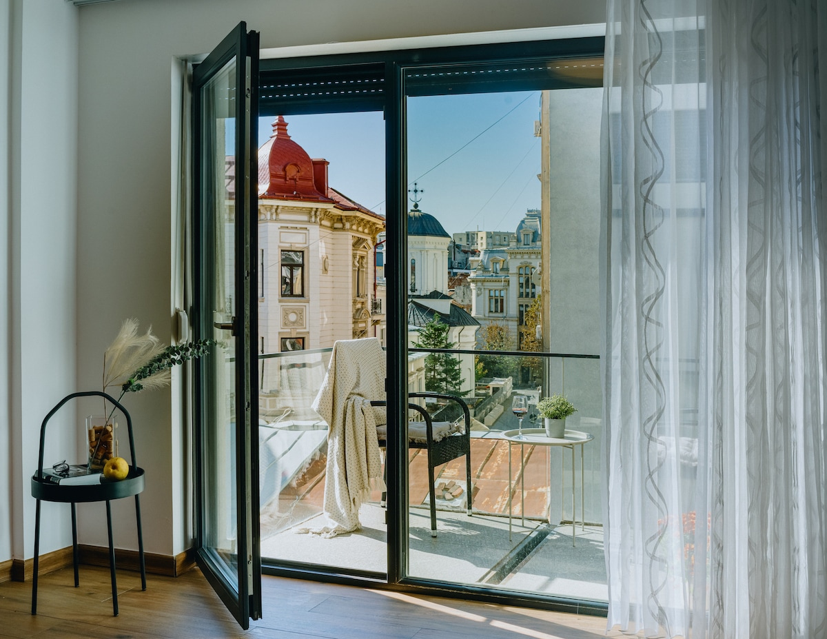 A balcony view displays charming architectural details from nearby buildings, framed by an open glass door. A cozy chair and a small table are positioned on the balcony, with a soft blanket draped casually over the chair. Natural light fills the interior space.